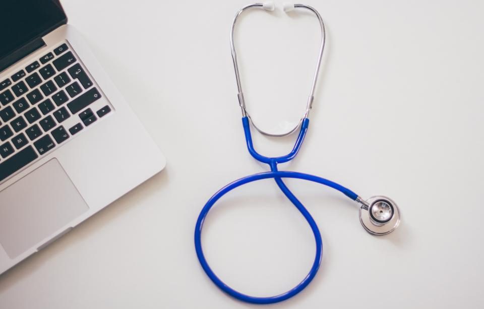 In a brief gap between patients, Han Do-gyeong turns the monitor to point at a patient's "past flow" — the desk holds only a blood-pressure cuff and one monitor
